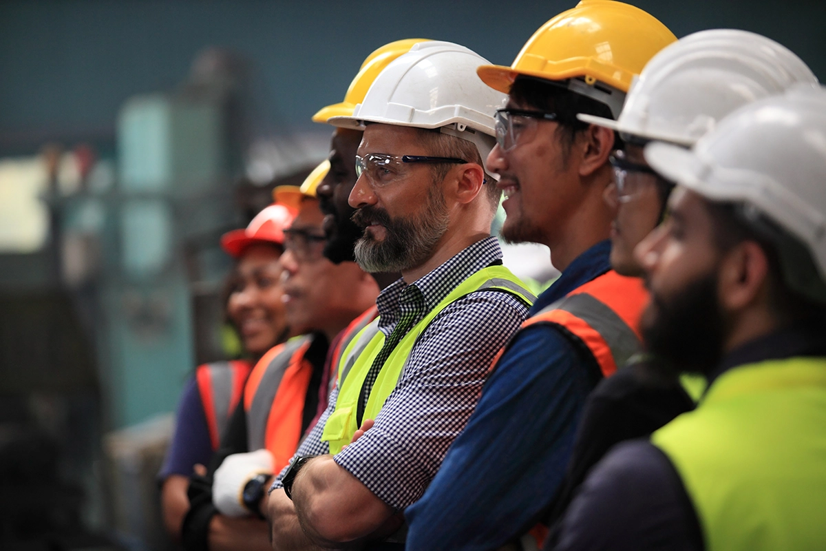 A group of construction workers wearing hard hats and safety goggles stands in a row, smiling.
