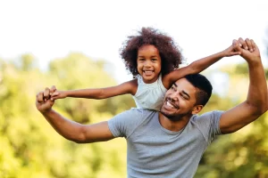 A father carries his smiling young daughter on his back.