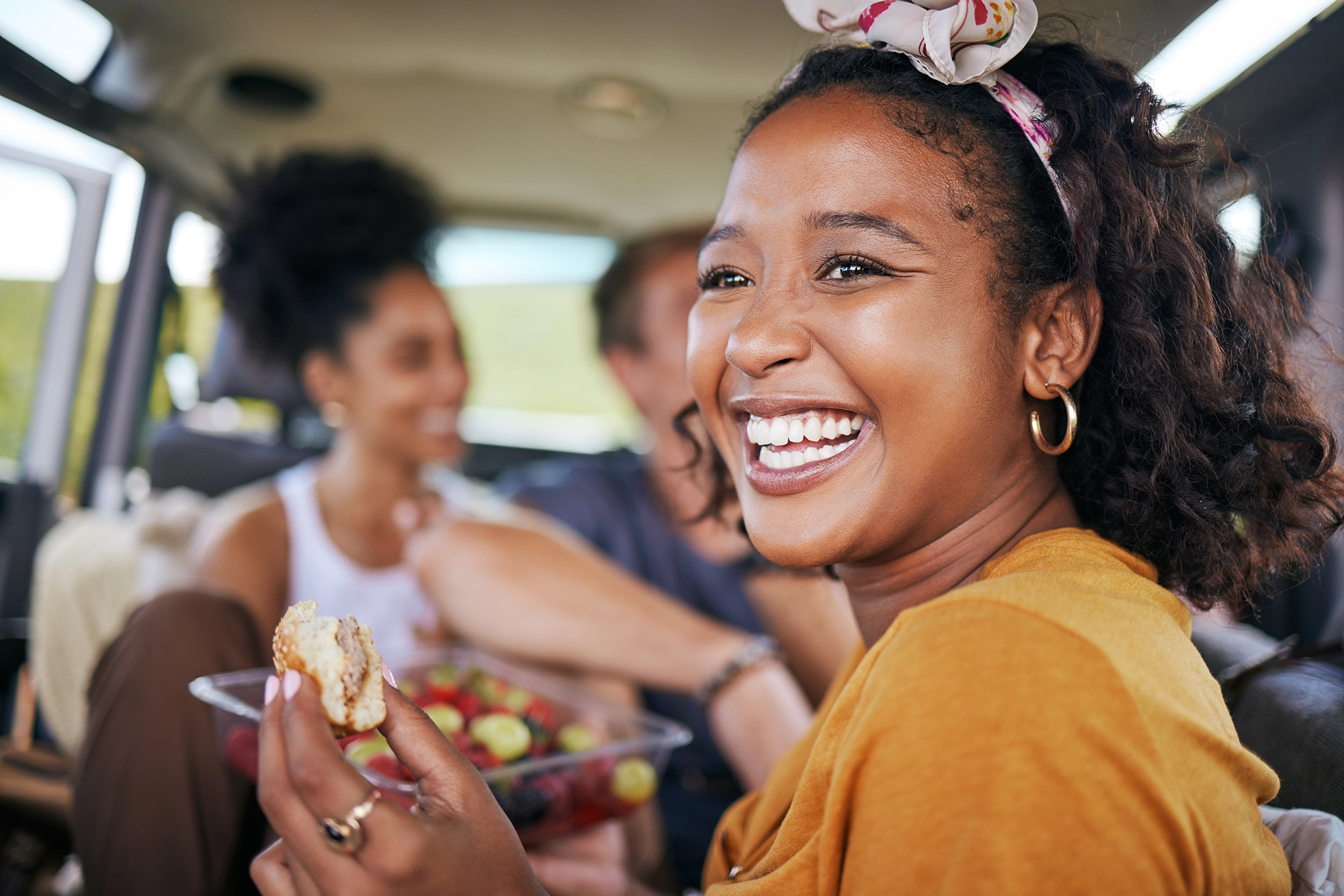 A woman wearing a yellow top sits in a car with friends, smiling and holding a piece of fruit.
